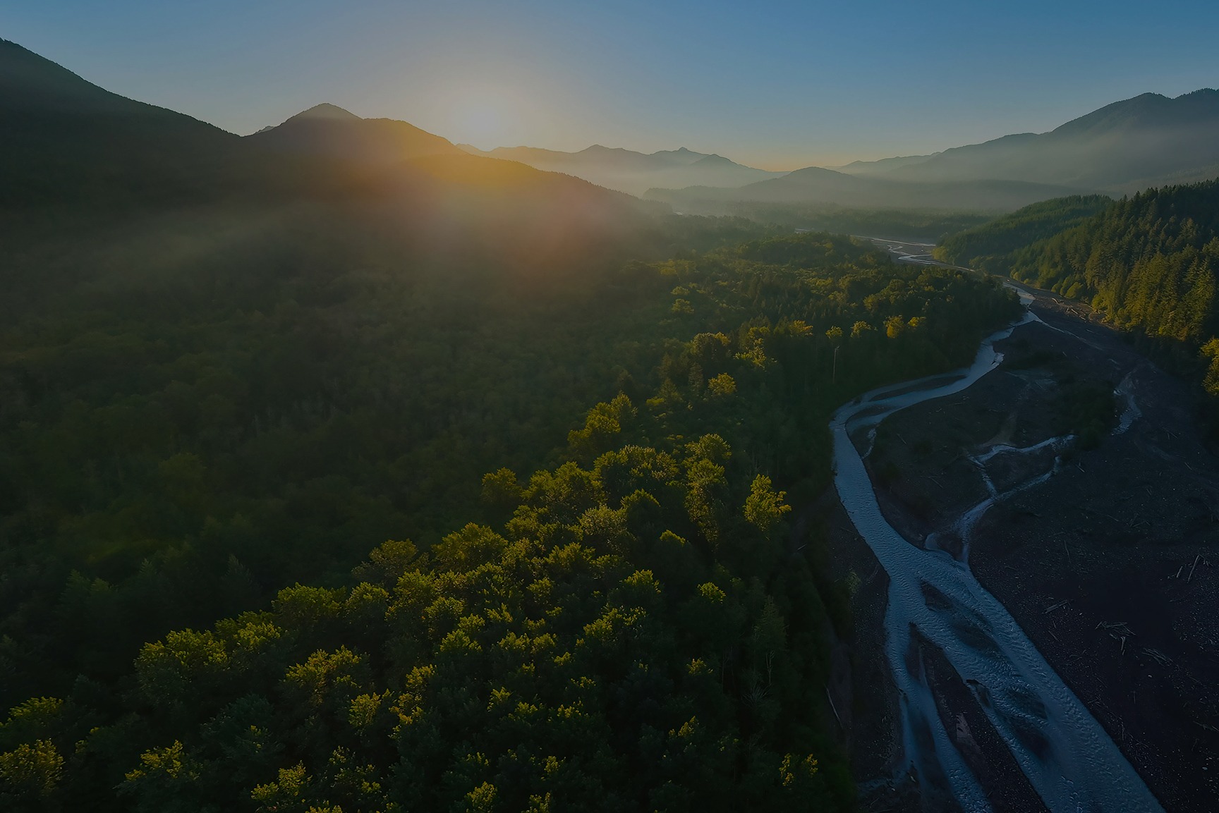 Aerial view of a winding river through a forested valley with mountains at sunset